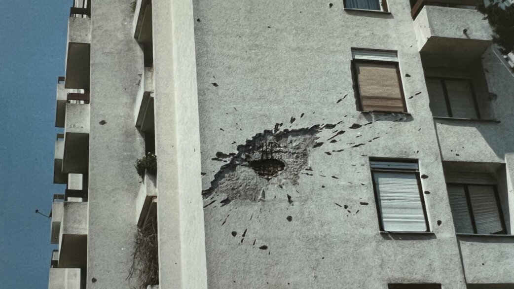 A grey concrete apartment building in Mostar showing significant war damage, with a large shell hole and shrapnel marks scattered across the facade between windows and balconies.