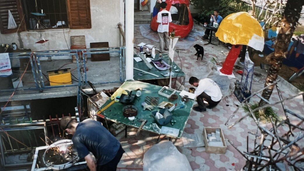 An outdoor courtyard workshop scene in Mostar showing several people creating colourful sculptures from recycled materials, including a red and yellow umbrella-like structure, with a black dog nearby.