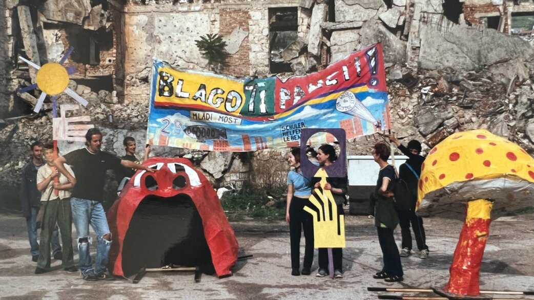 A group of people display colourful handmade sculptures and a banner reading "Blago ti Fansi" amid war-damaged buildings in Mostar, Bosnia Herzegovina. Sculptures include a red mouth-shaped form and spotted yellow mushroom.
