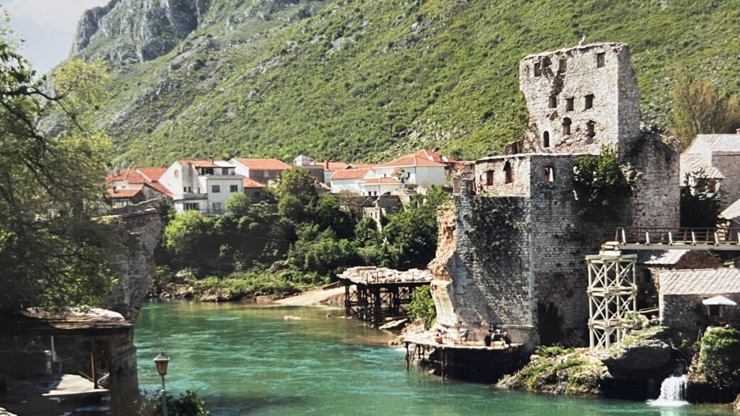 Photograph of Mostar during post-war reconstruction, showing the destroyed Stari Most bridge site with its remaining stone towers, the turquoise Neretva River, and hillside buildings.