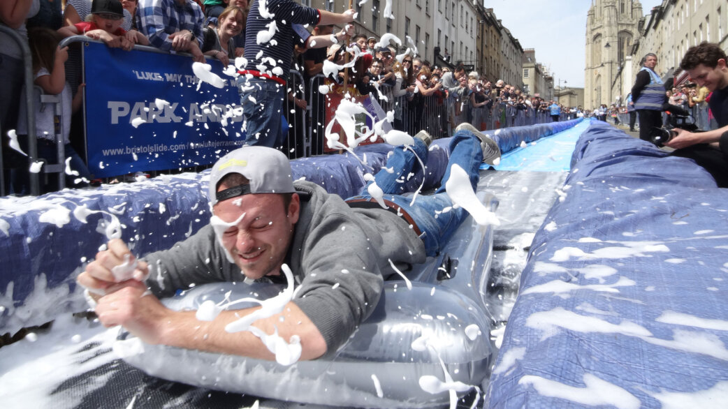 A man on an inflatable lilo slides down a long blue water slide on Park Street, Bristol, surrounded by splashing foam and cheering spectators lining both sides.