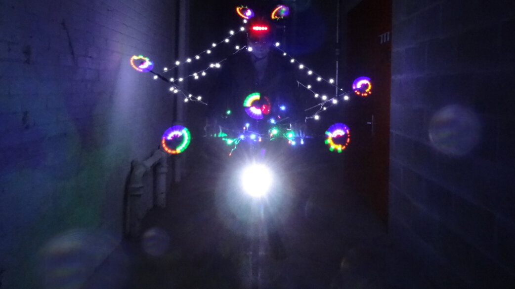 A cyclist rides through a dark alleyway at night, their bicycle adorned with colourful LED lights forming circular patterns and string lights creating a diamond shape around them.