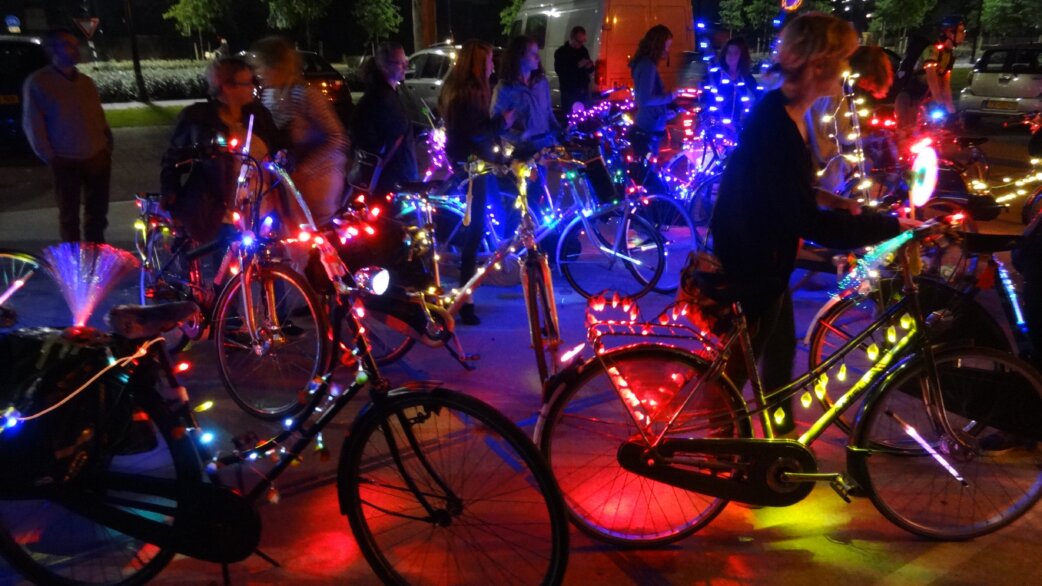 A nighttime gathering of cyclists with bicycles decorated in colourful LED fairy lights, creating a festive, glowing atmosphere in an urban setting.
