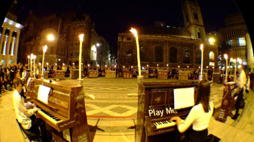 A nighttime fisheye photograph showing 21 pianos arranged in a circle outside London's Guildhall, each lit by tall candles, with pianists performing and crowds watching.