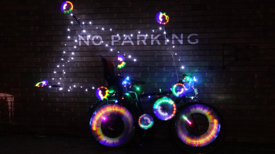 A bicycle decorated with colourful LED lights on its wheels and frame, parked at night against a brick wall with "NO PARKING" painted on it.