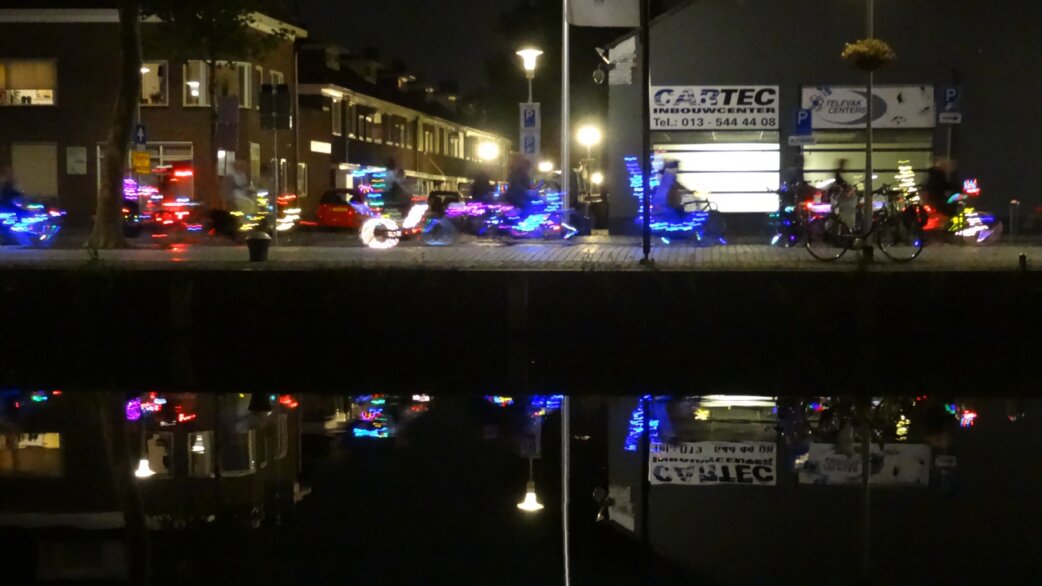Nighttime photograph of cyclists with LED-lit bicycles riding along a canal in the Netherlands, their colourful lights reflecting in the dark water below.