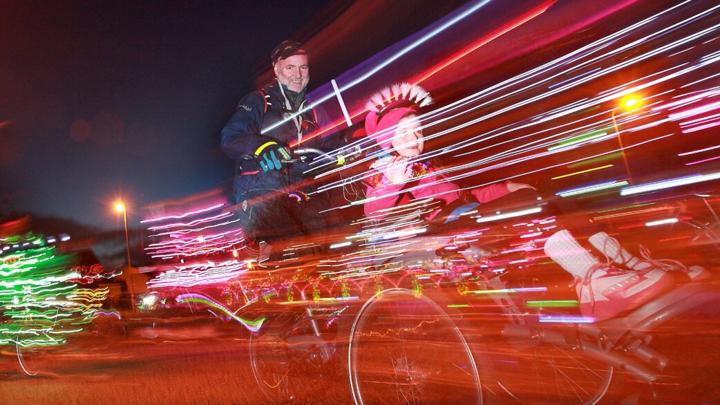 A man and child cycling at night through colourful light trails, the child wearing a festive headdress. Long exposure creates streaks of red, blue and white light.