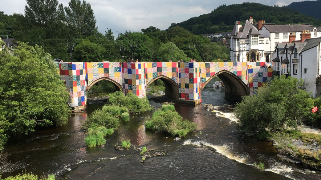A medieval stone bridge with three arches spanning a river, wrapped entirely in colourful patchwork fabric squares. Welsh town buildings and green hills visible in the background.