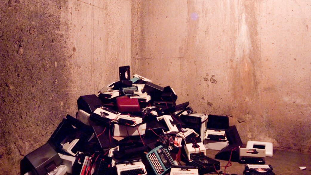 A pile of abandoned typewriters in various colours heaped in the corner of a dimly lit concrete tunnel, creating an atmosphere of industrial decay and obsolescence.
