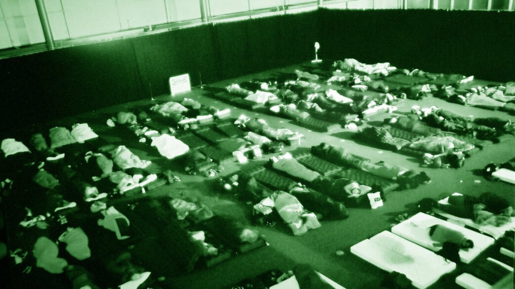 Night-vision photograph showing dozens of people lying on mattresses and bedding spread across a large indoor floor space, appearing to sleep during a group event.