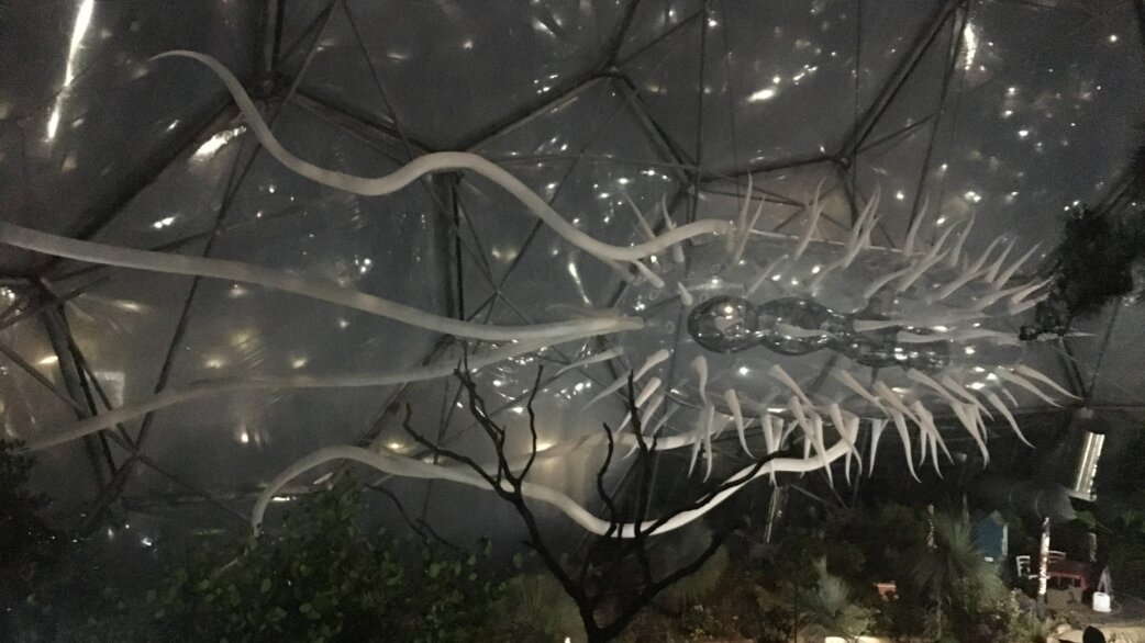 A 30-metre translucent inflatable E.coli sculpture suspended inside a geodesic dome, featuring elongated flagella and numerous hair-like pili, illuminated against the dark structure.