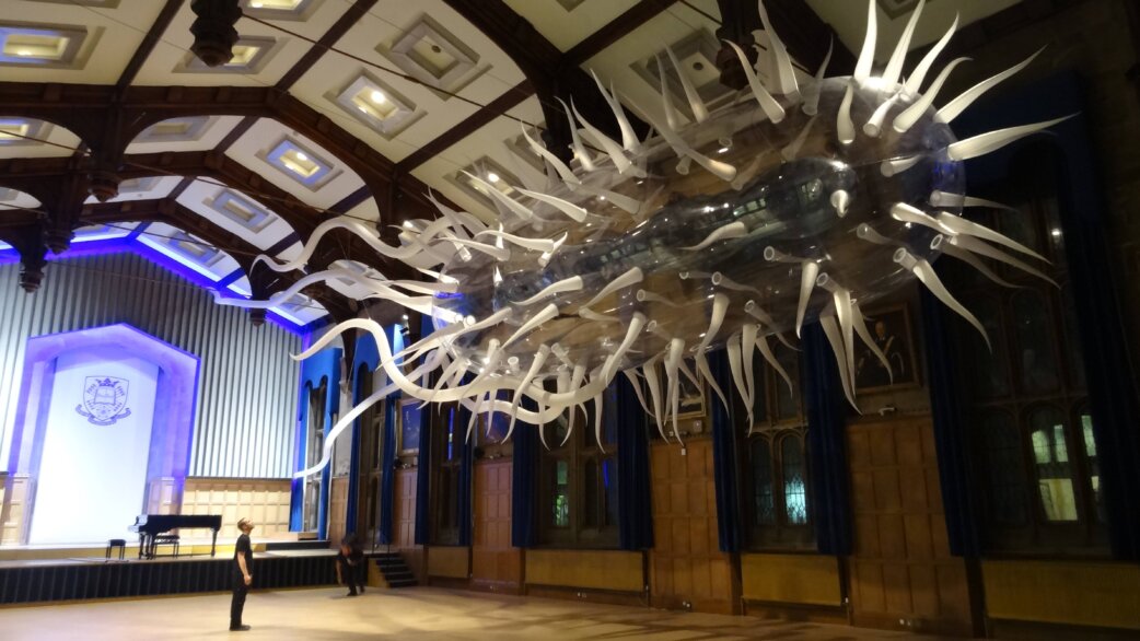 A 30-metre inflatable E.coli sculpture with white tentacle-like flagella suspended in Firth Hall, Sheffield, dwarfing a person standing beneath its translucent body.