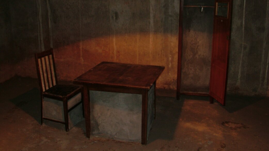 A wooden chair and table appear partially encased in concrete against stained grey walls, with an open red cabinet nearby, creating an eerie abandoned atmosphere.