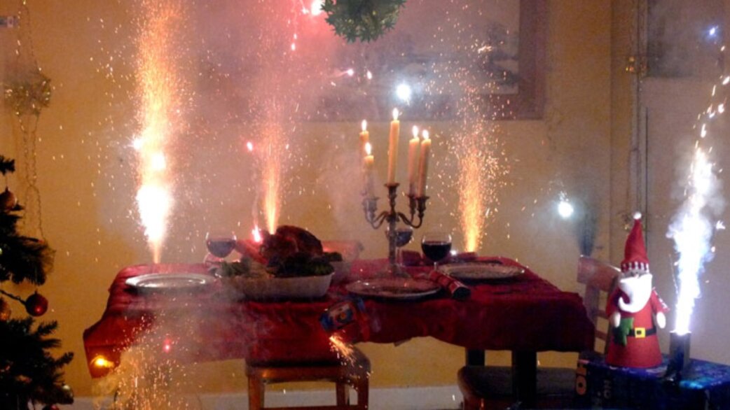 A Christmas dinner table with lit candles and indoor fireworks erupting beside a roast turkey, wine glasses and place settings, with a Santa figure and Christmas tree visible.