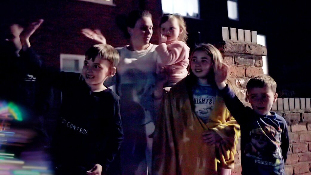 A family group including a woman holding a toddler, three boys and a teenage girl stand outside at night, waving and looking upward with colourful light reflections visible.