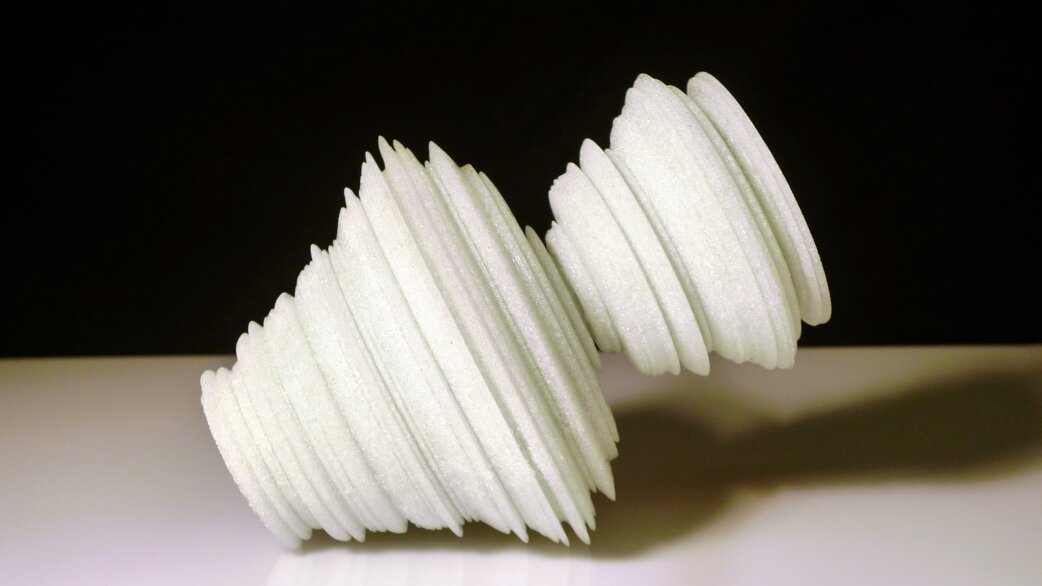 A white glass sculpture resembling two conical spiral forms joined at their points, made from stacked circular discs of varying diameters, approximately 21cm long.