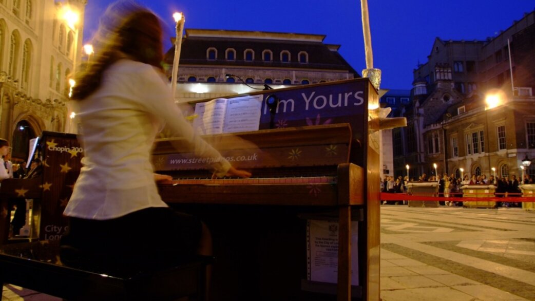 A pianist in a white jacket plays a candlelit street piano at dusk in Guildhall Yard, London, with audience members visible behind barriers in the background.