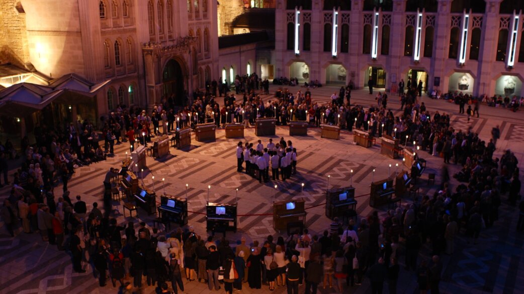 An aerial view of 21 pianos arranged in a circle with candles, performers in white at the centre, surrounded by spectators in the Guildhall courtyard at night.