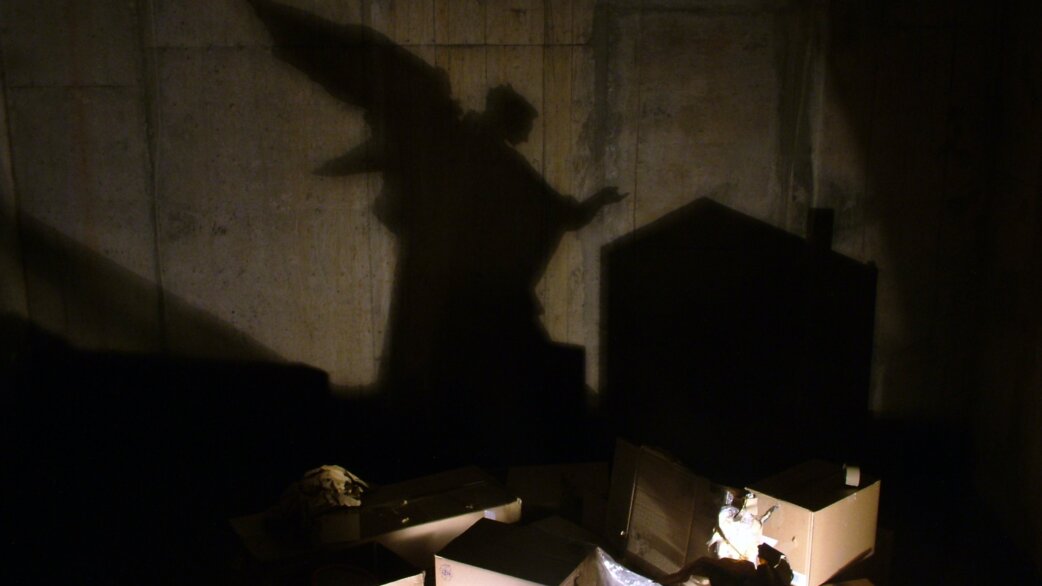 A dramatic shadow of an angel with outstretched wings projected onto a concrete tunnel wall, cast by scattered mail, bank statements and debris illuminated below.