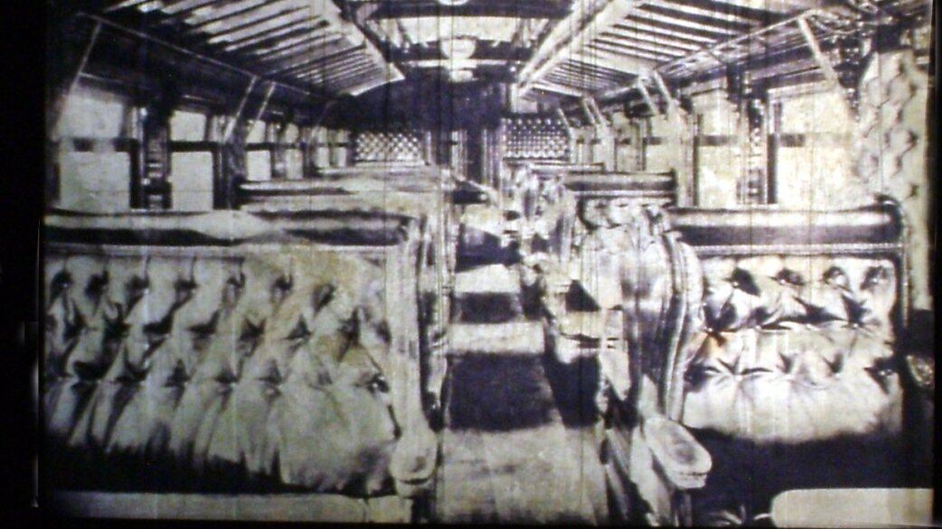 A high-contrast projected image showing the interior of an ornate Victorian train carriage with plush tufted seating, displayed within a dark tunnel space.