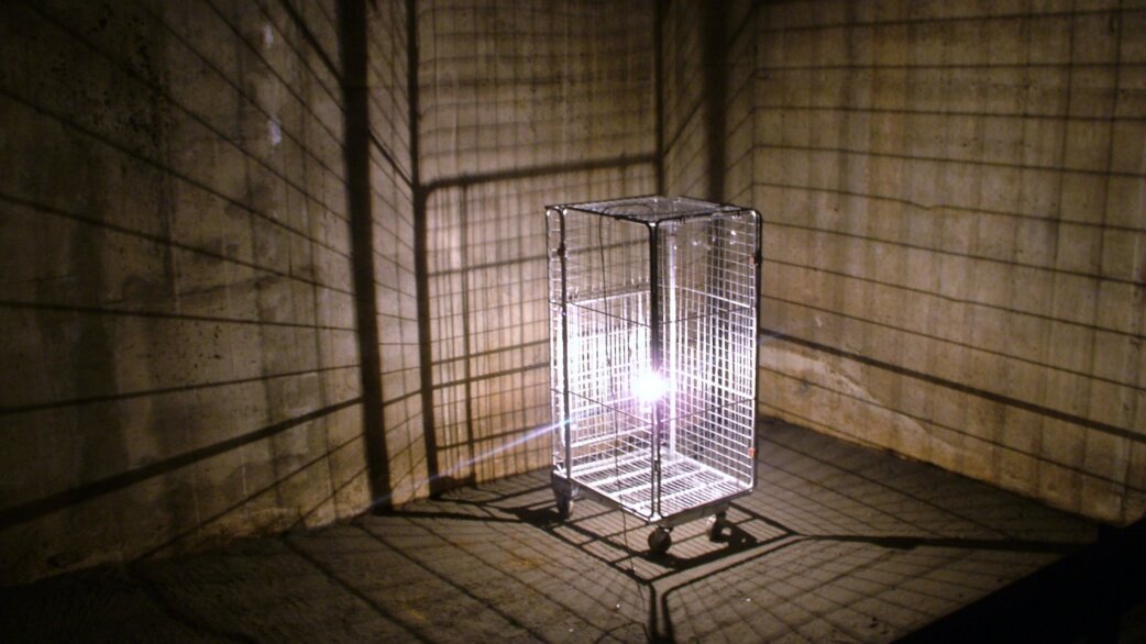 A wire mesh industrial cage on wheels with a bright light inside, casting grid-like shadows across concrete tunnel walls in a dark underground space.