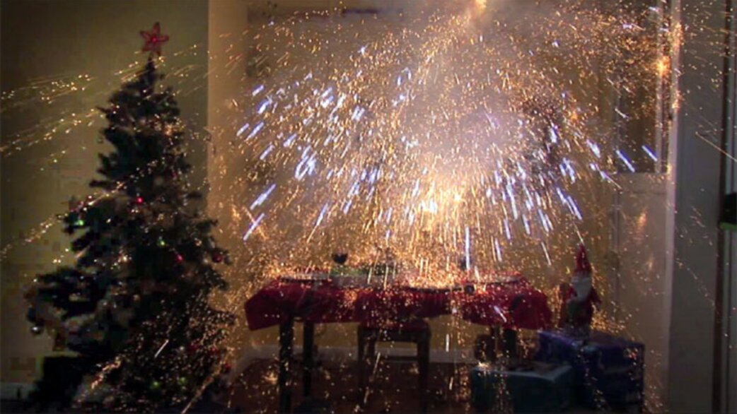 Indoor scene showing fireworks or sparklers exploding dramatically beside a decorated Christmas tree, with golden and white sparks filling a living room space.