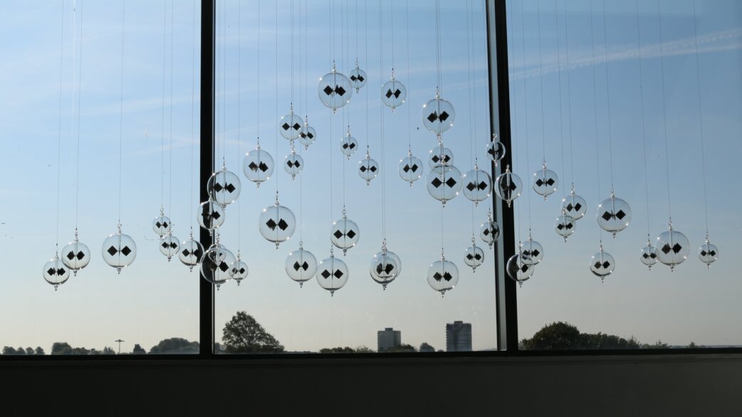Glass radiometers suspended on fine wires in front of large windows, forming a chandelier installation. Each spherical radiometer contains black and white vanes. Trees and sky visible beyond.