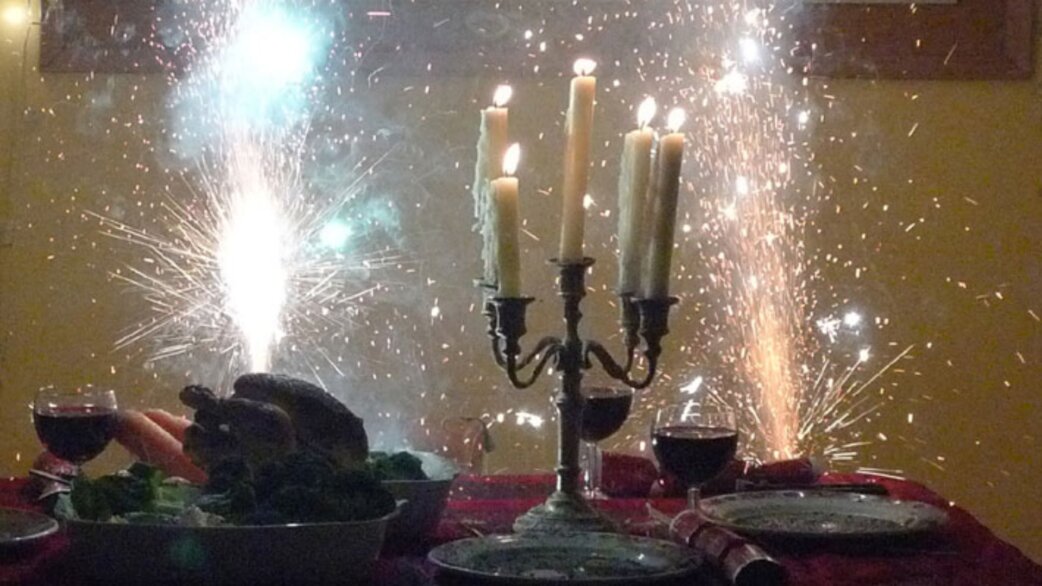 A festive dinner table with lit candelabra, glasses of red wine, salad bowl and roasted meat, flanked by two indoor sparkler fireworks showering bright sparks.