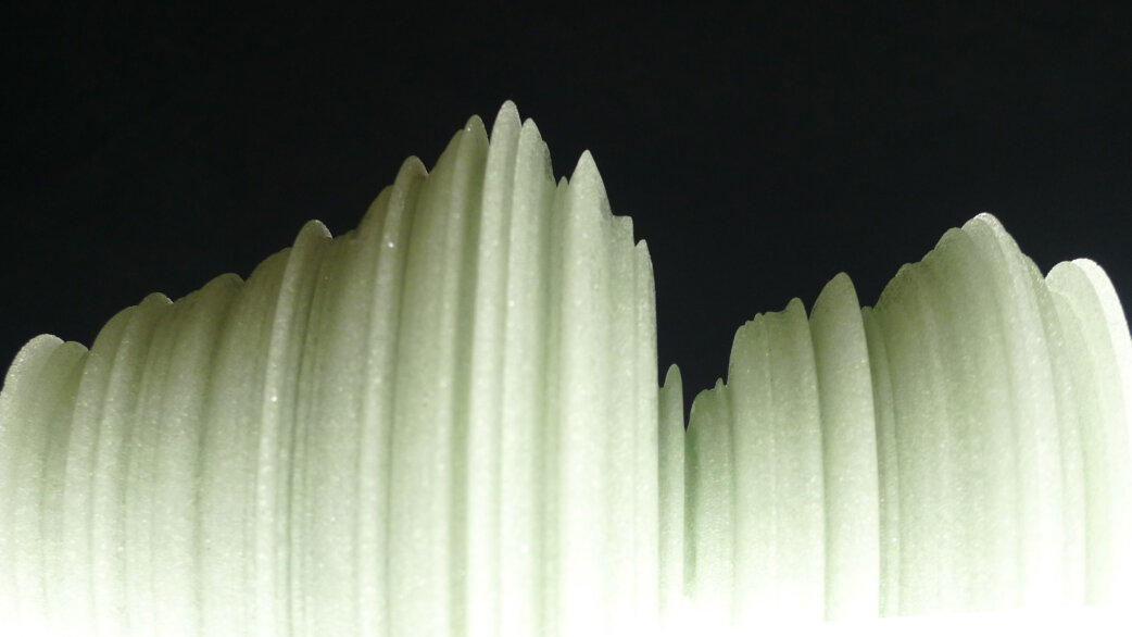 A translucent pale green glass sculpture with vertical ridges forming peaks and valleys, resembling a mountain range, set against a black background. The form visualises stock exchange data.