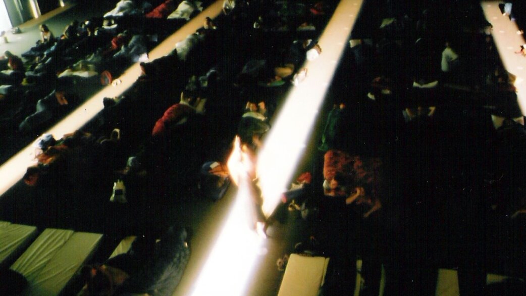 An audience lies on reclining seats in a darkened auditorium, viewed from above. Light streaks cross the image, and people appear to be resting or sleeping during a performance.