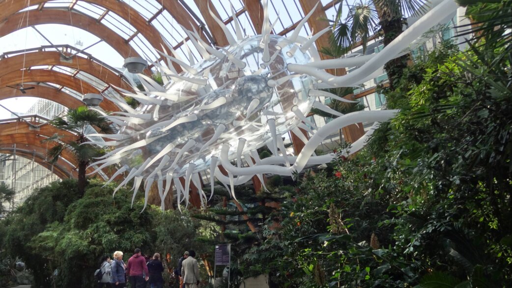 A 30-metre white inflatable E.coli sculpture with elongated flagella suspended inside Sheffield Winter Gardens' glass atrium, surrounded by tropical plants and visitors below.