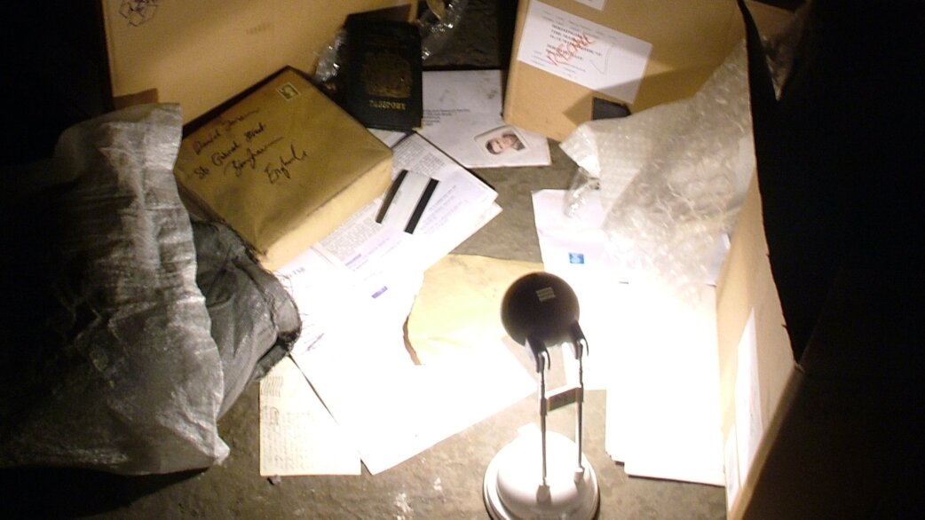 A dimly lit scene of scattered documents, passports, envelopes, bubble wrap and bank statements illuminated by a desk lamp, forming a crime scene installation in an abandoned tunnel.