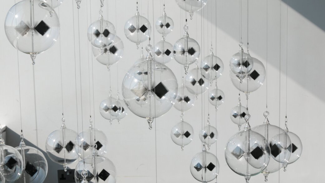 Dozens of clear glass radiometers suspended on thin wires against a white wall, each containing black and white vanes, catching sunlight and casting soft shadows.
