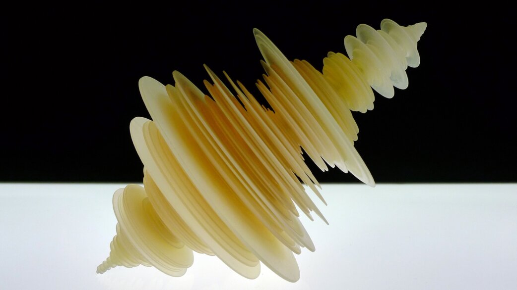A translucent pale yellow 3D-printed sculpture resembling layered, rotating discs that spiral outward, created from seismogram data of the 2011 Tōhoku earthquake, approximately 30cm wide.