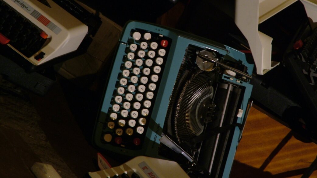 A pile of abandoned vintage typewriters in blue, cream and white, lying in darkness. The central turquoise typewriter has round white keys and visible typebars.