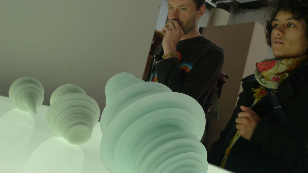 Two visitors contemplate illuminated glass sculptures resembling stacked spherical forms, approximately 20-30cm tall, displayed on a light table. The translucent pieces visualise stock exchange data.