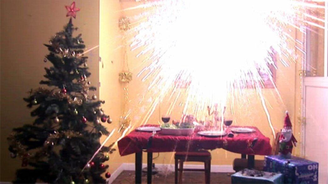 A decorated Christmas tree beside a dining table set for dinner, with a firework exploding indoors sending bright sparks across the room.