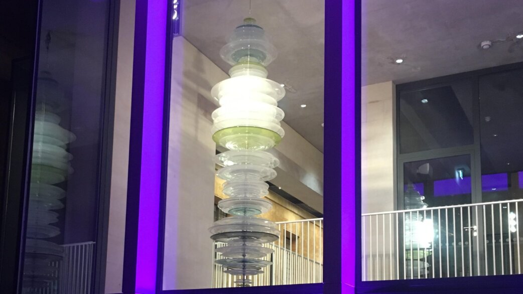 A tall vertical glass sculpture made of approximately 80 stacked hand-blown glass discs in muted greens and clear tones, illuminated and suspended in a modern building foyer with purple-lit window frames.
