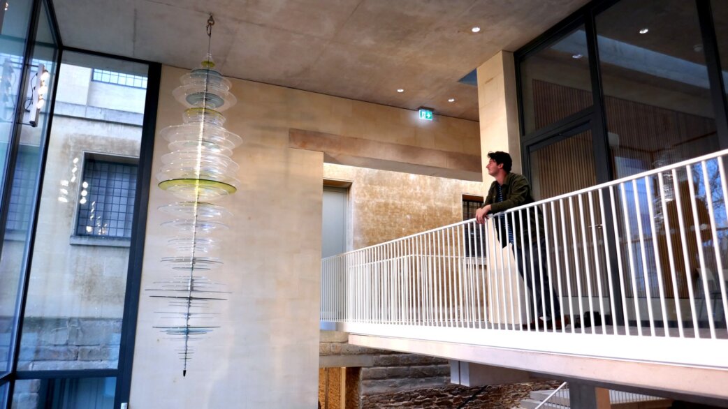 A tall glass sculpture of stacked circular discs in clear and pale green tones hangs in a modern foyer, approximately 3.6 metres long, with a person viewing from a balcony.