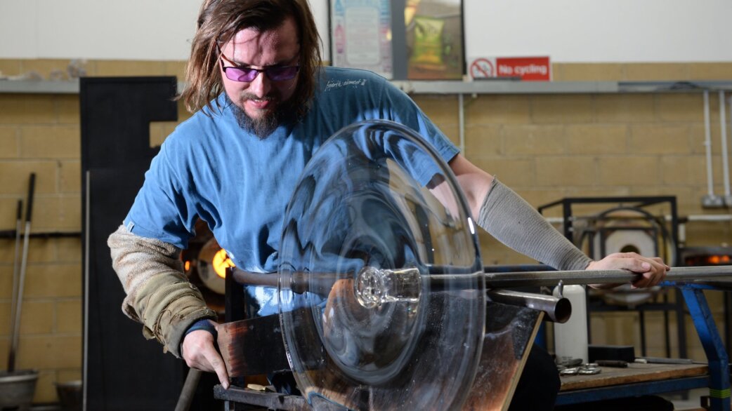 A glassblower in protective glasses and arm guard shapes a large clear glass roundel, approximately 60cm in diameter, attached to a metal blowpipe in a workshop.