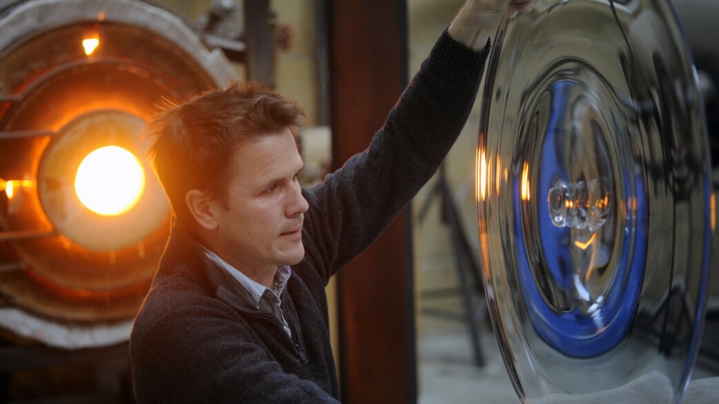 A man in a dark jumper examines a large glass sculpture made of concentric circular roundels with blue and clear glass, beside a glowing furnace in a glassblowing workshop.