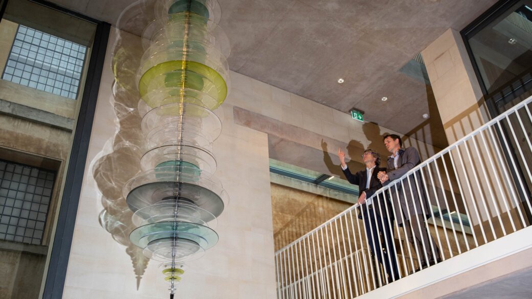 A tall glass sculpture of stacked coloured roundels, approximately 3.6 metres long, hangs in a building foyer as two people observe from a staircase balcony.