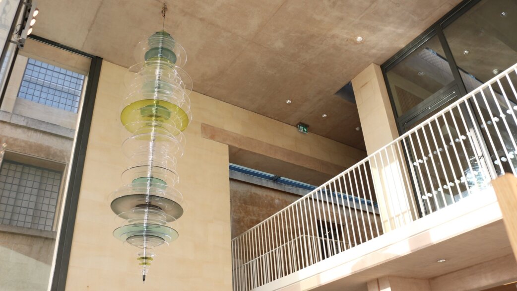 A vertical glass sculpture made of approximately 80 hand-blown coloured glass discs in greens, yellows and clear tones, suspended in a modern building foyer, measuring 3.6 metres in length.