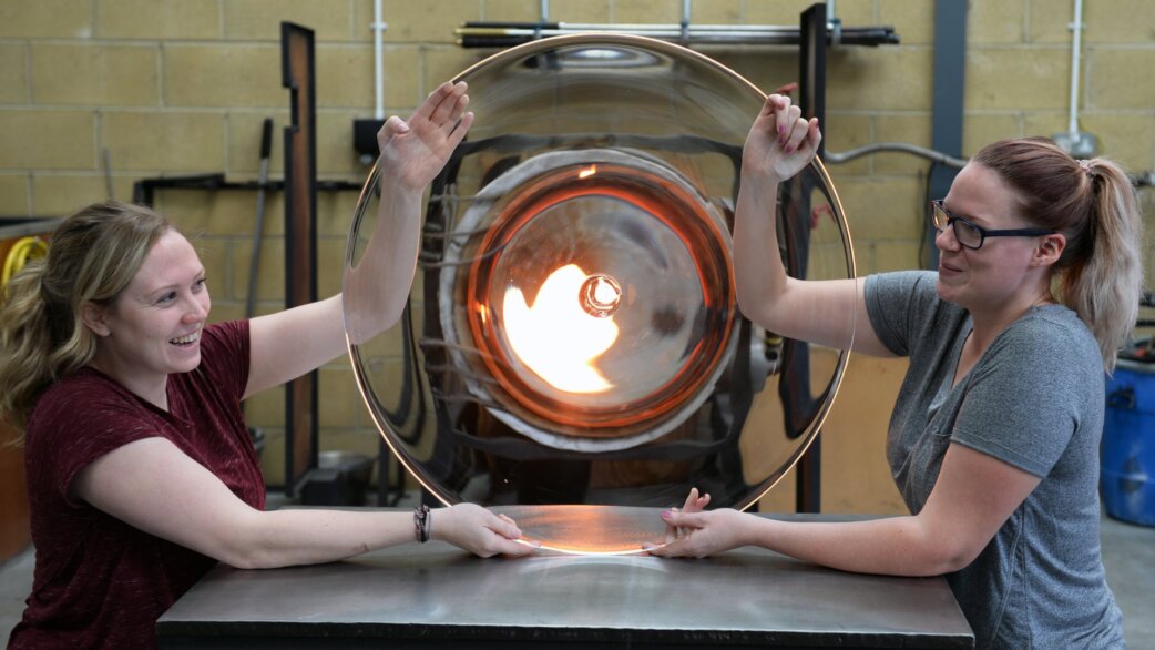 Two women hold up a large circular glass roundel, approximately one metre in diameter, in front of a glowing furnace in a glassblowing workshop.