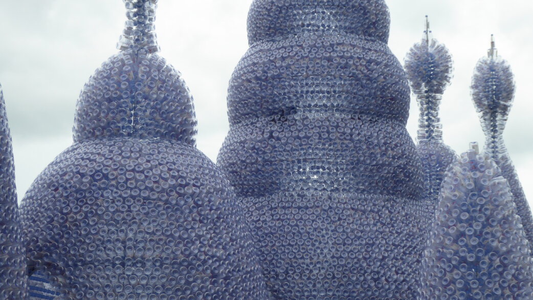 Sculptural towers resembling bulbous, organic forms constructed entirely from thousands of clear plastic water bottles, creating a translucent blue-tinged surface against an overcast sky.