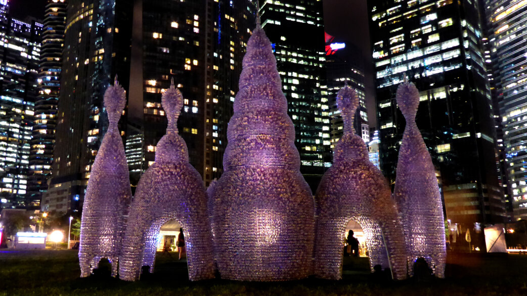 Illuminated sculptural pavilion made from 25,000 plastic bottles forming spired towers resembling radiolarians, approximately 8 meters tall, displayed at night against Singapore's skyline.