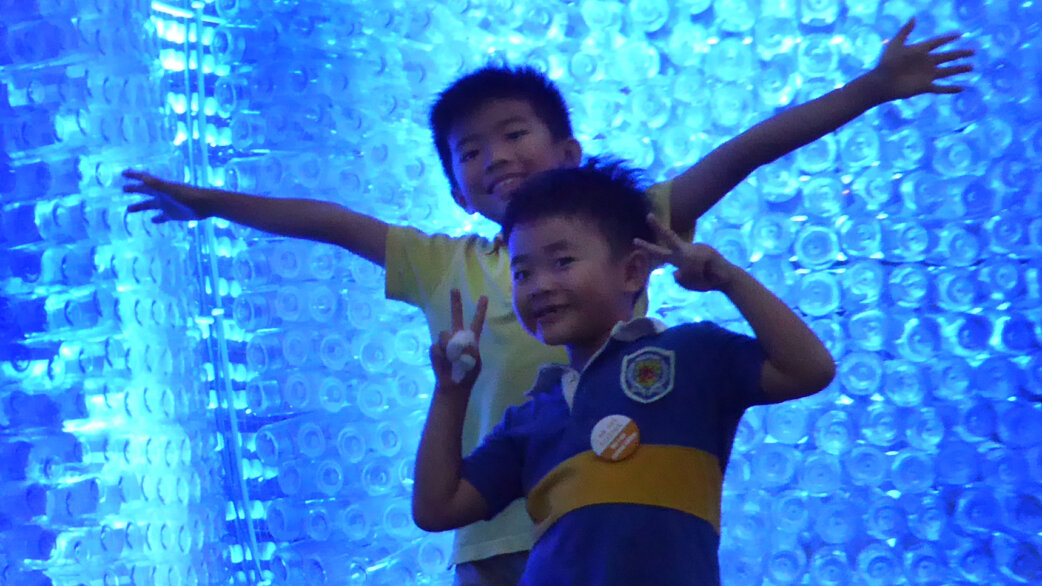 Two children posing playfully inside a glowing blue pavilion structure made from thousands of plastic water bottles, with one making peace signs and the other with arms outstretched.