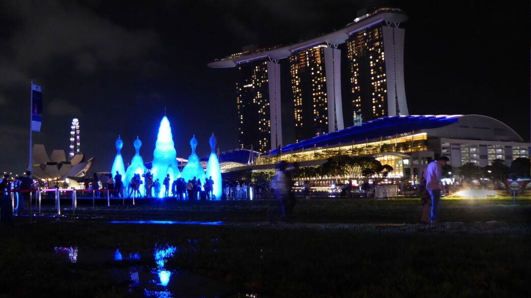 Nighttime view of Ocean Pavilion, illuminated blue sculpture made from 25,000 plastic bottles shaped like radiolarians, with Marina Bay Sands hotel and crowds in background, Singapore.