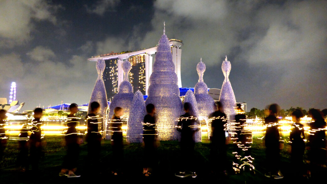 Nighttime view of illuminated spire-shaped sculptures made from plastic bottles, resembling radiolarians. A long exposure gives horizontal streaks of light and blurred visitors in foreground.