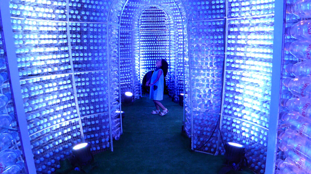 A person stands inside a glowing blue tunnel structure made from thousands of plastic water bottles arranged in arched walls, illuminated from within, creating an immersive radiolarian-inspired pavilion.